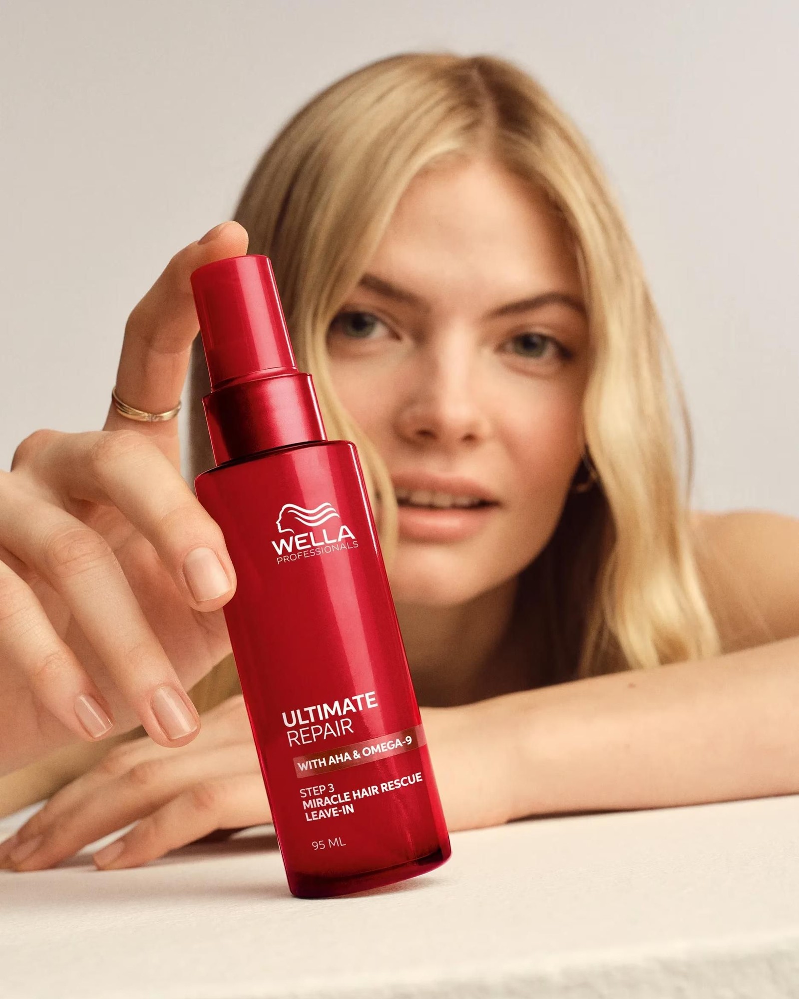 Blonde woman holding a red Wella Ultimate Repair Miracle Hair Rescue Leave-In bottle toward the camera