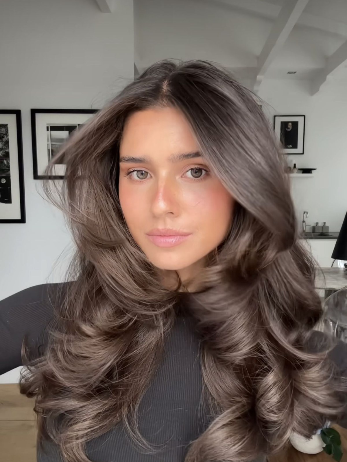 Woman with long brunette bouncy curls swept off the face, shown from the shoulders up in a bright room with framed photographs