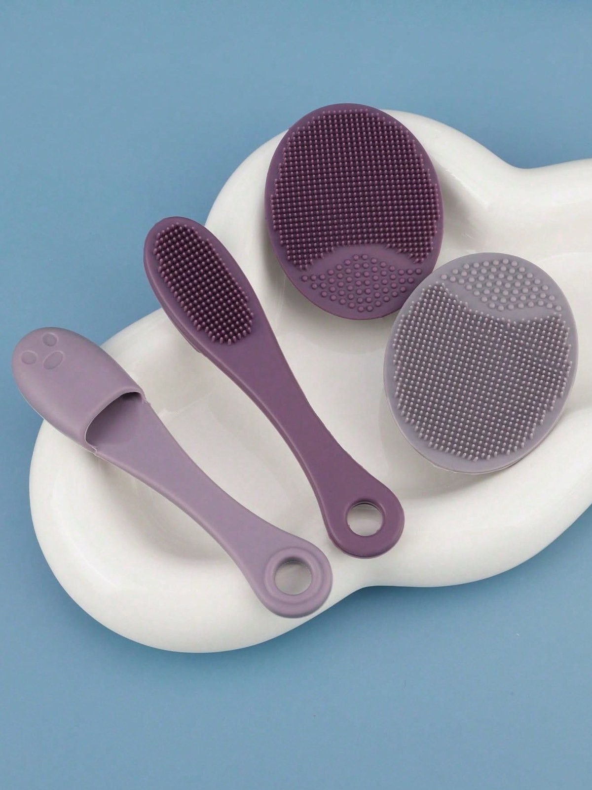 Purple and gray silicone body brushes and scrubbers arranged on a white ceramic dish against a blue background