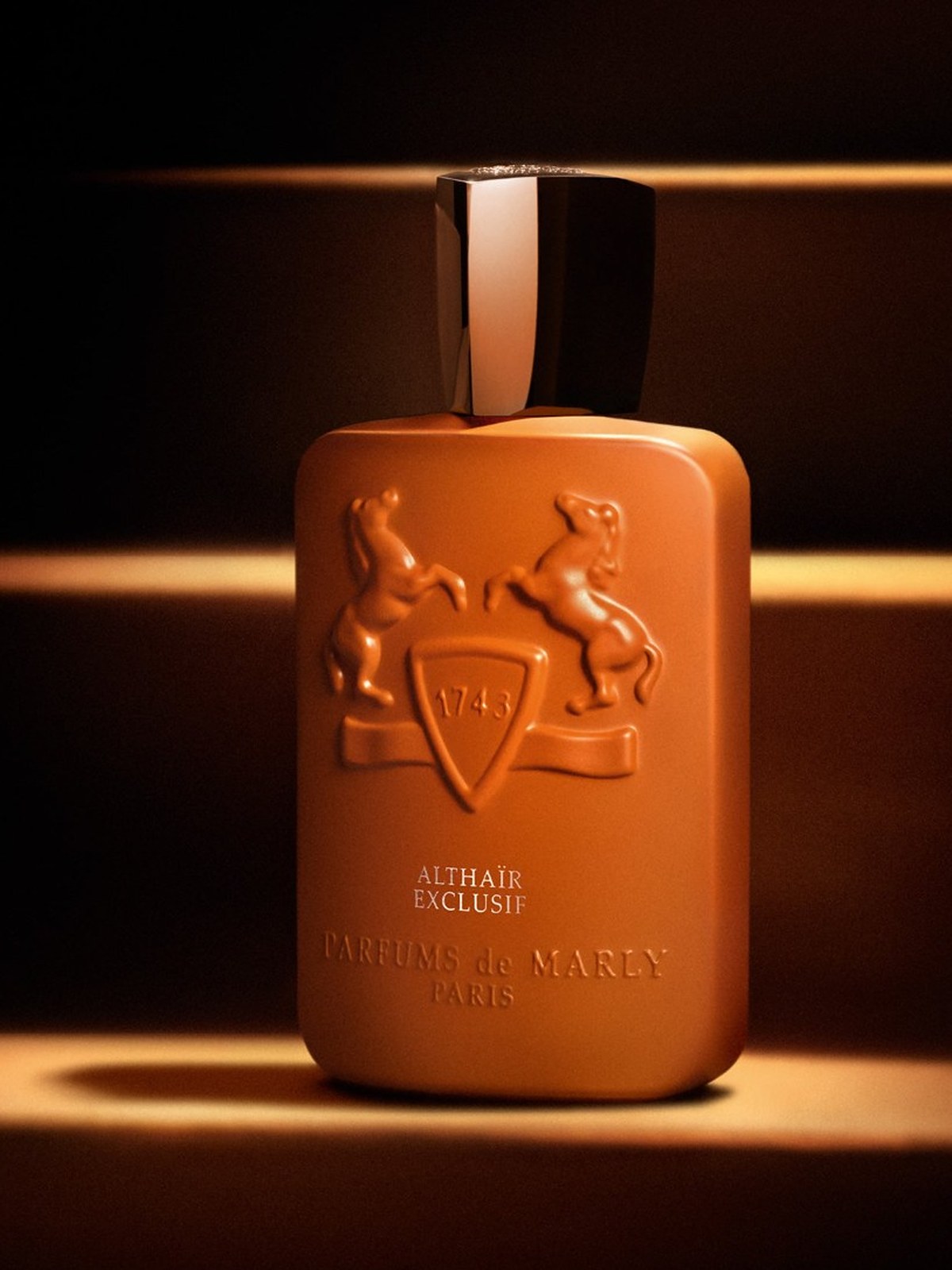 Orange Parfums de Marly bottle lit in horizontal bands of light on a dark ground