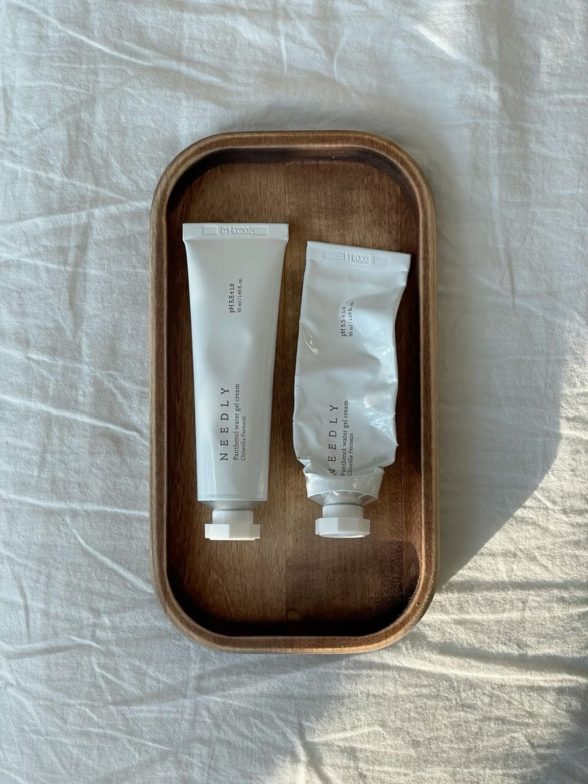 Two white NEEDLY panthenol water gel cream tubes, one new and one squeezed flat, on a wooden tray over rumpled linen.