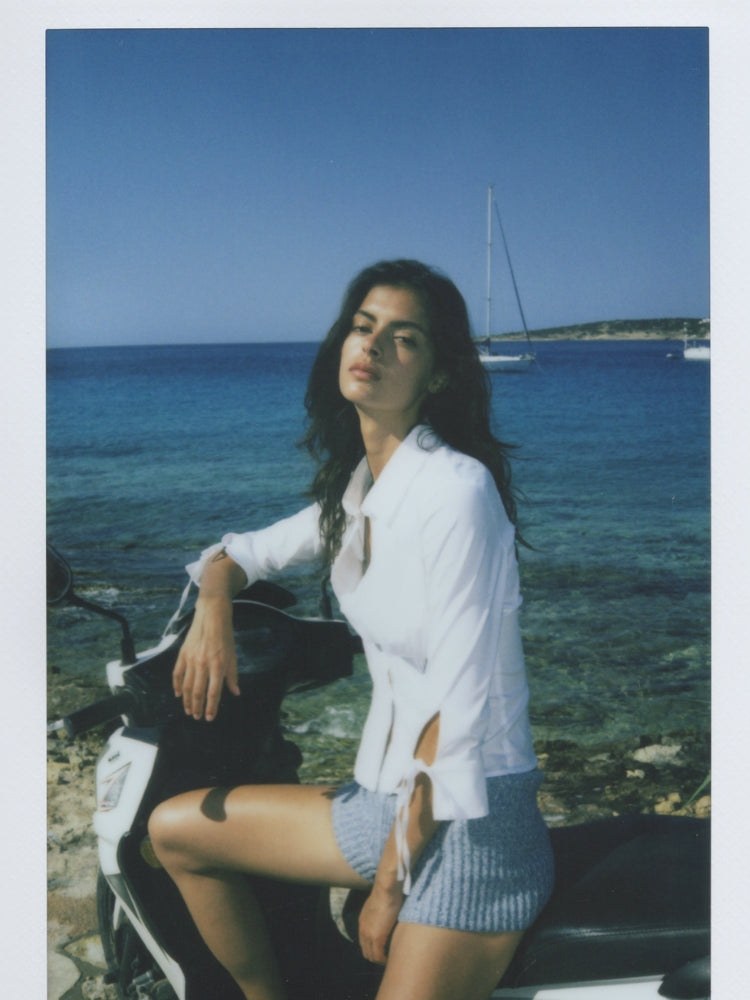 Model seated on a scooter by the sea in a white blouse and blue shorts