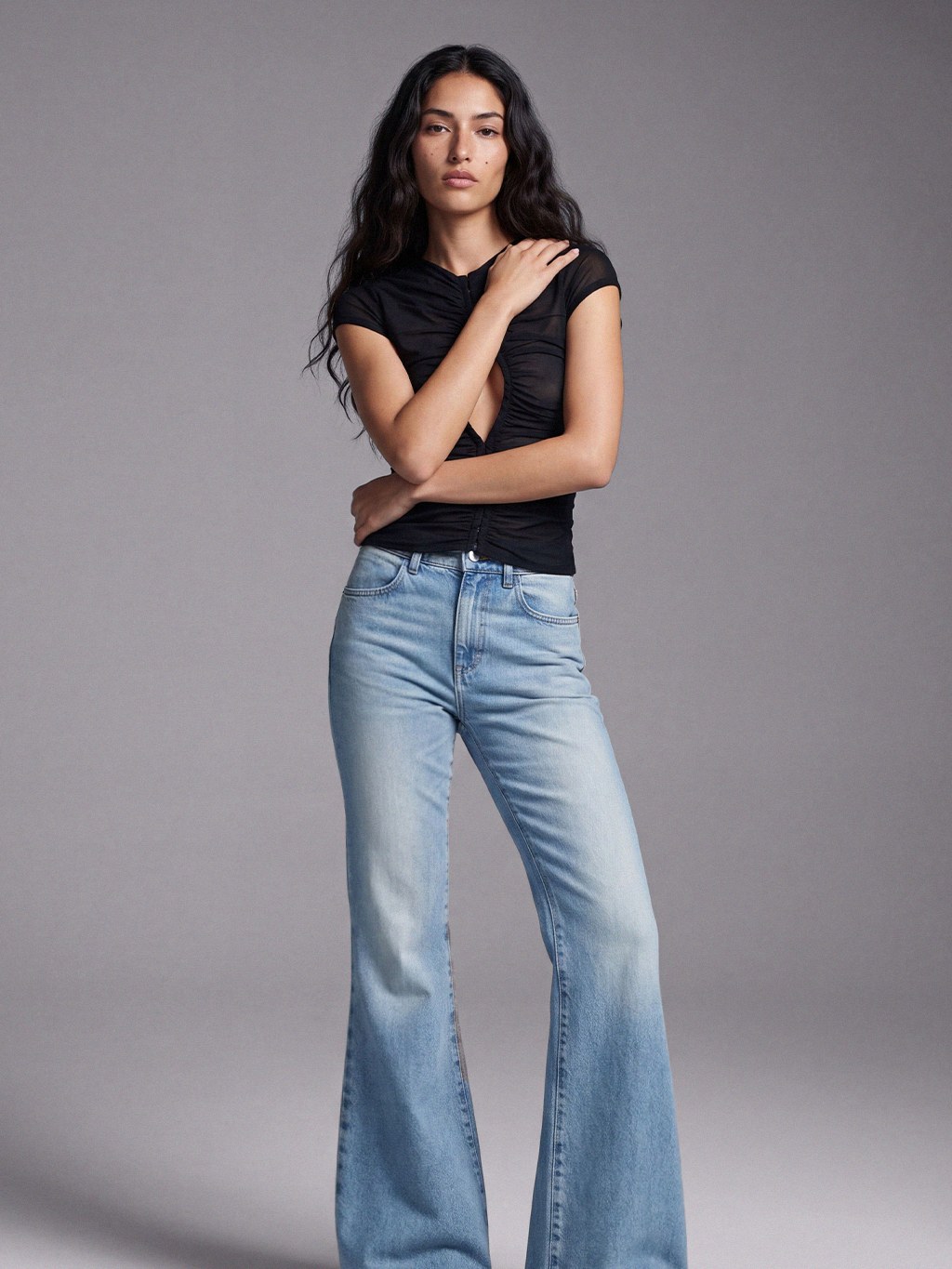 Model in flared light-wash jeans and a black top against a gray studio backdrop.