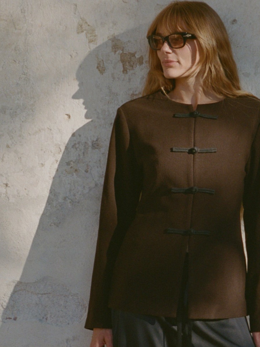 Model wearing a brown Ciao Lucia coat against a sunlit stone wall