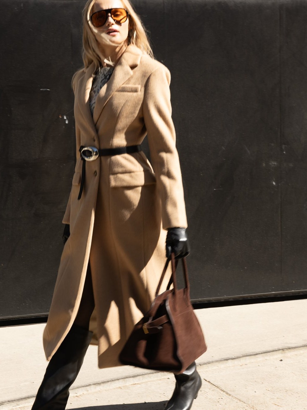 Woman in a long tan coat and sunglasses walking with a dark handbag