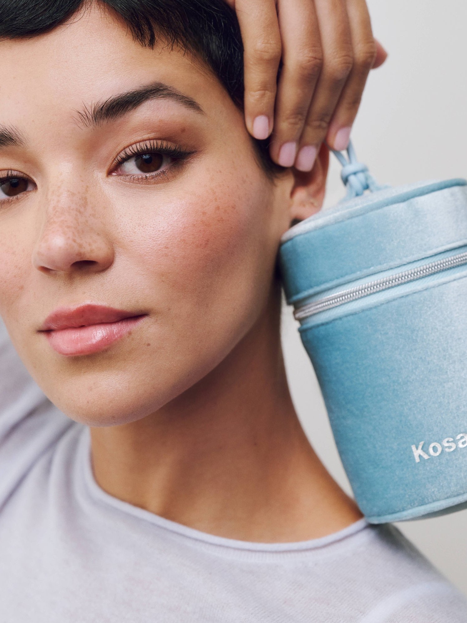 Woman with short dark hair holding a blue velvet Kosas makeup case by her face