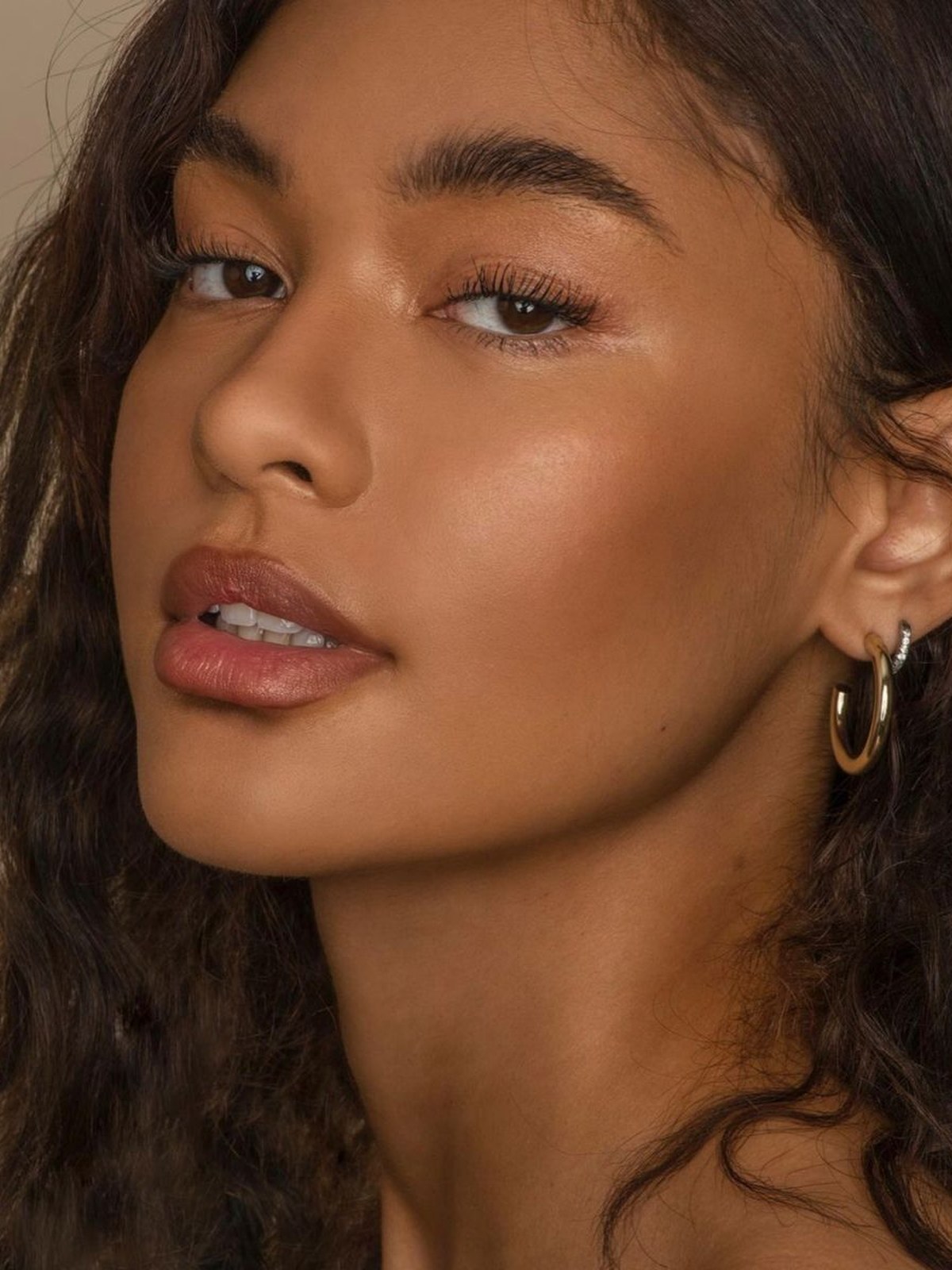 Close-up of a woman with wavy dark hair, glowing skin, a warm brown lip and gold hoop earrings