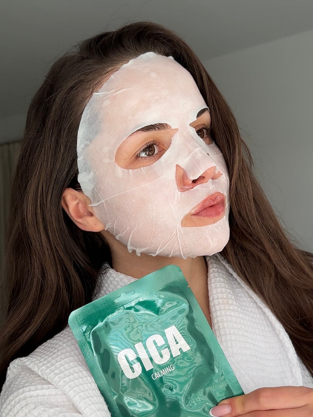 Woman in a white robe wearing a sheet mask and holding a green LAPCOS mask packet.
