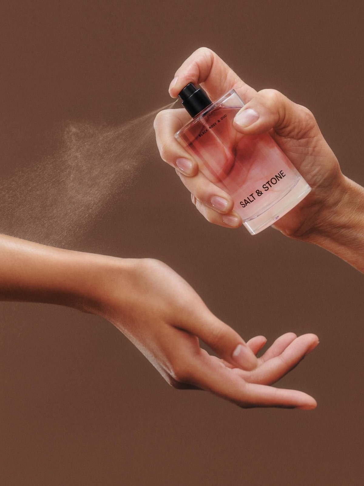 Hand spraying a Salt & Stone perfume bottle over an outstretched wrist against a warm brown backdrop