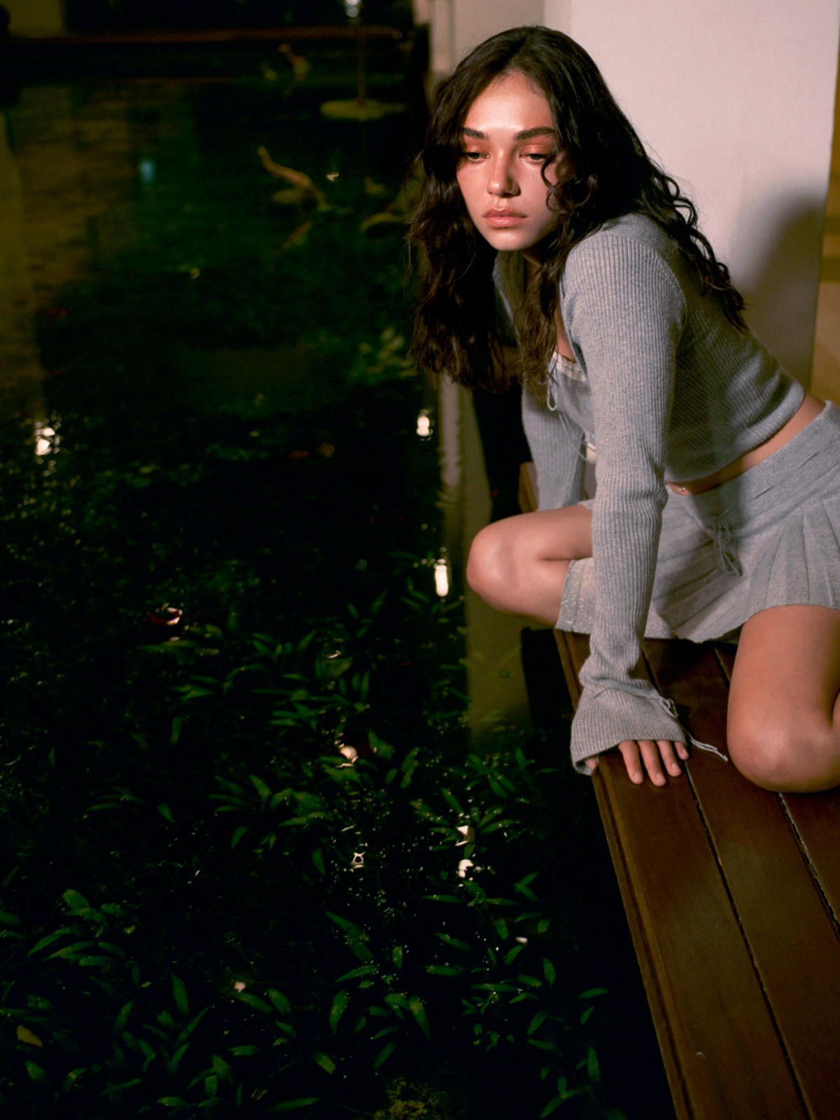 Model in a grey GUIZIO top and skirt seated beside greenery in a dim interior