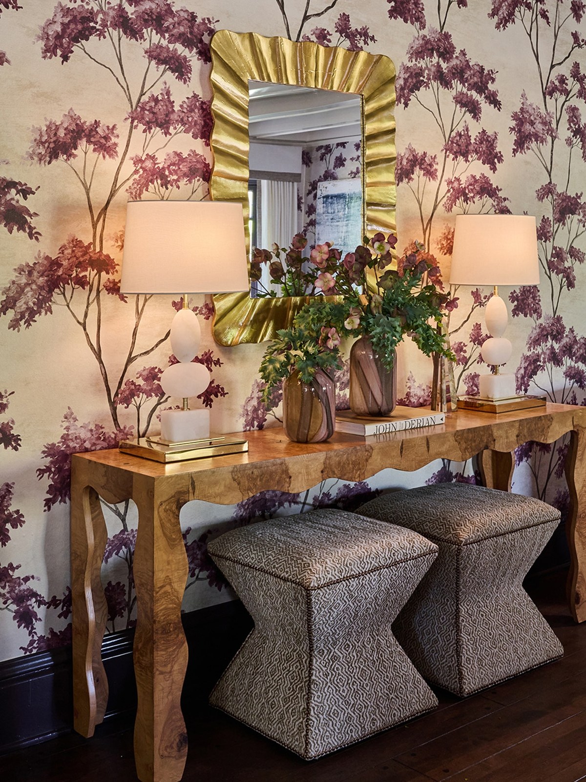 Wallpapered entry with a burl wood console, two table lamps and a gilt mirror