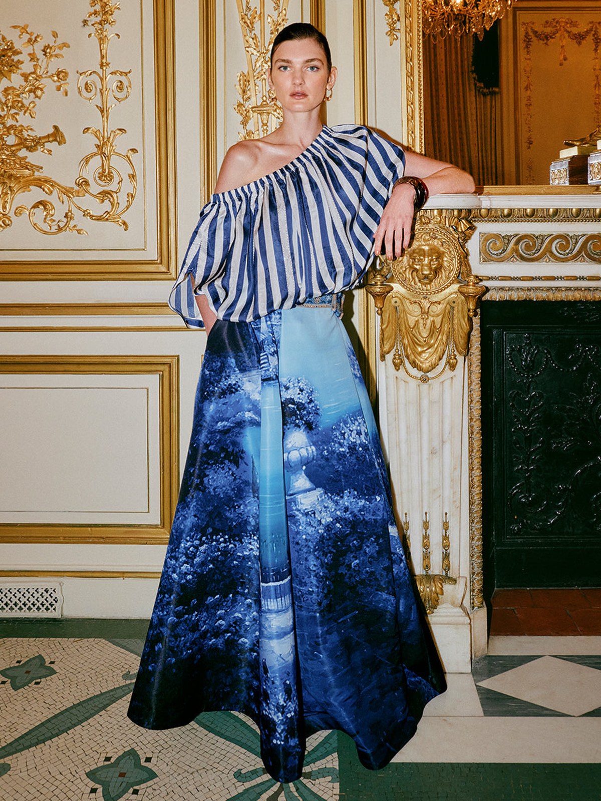 Model in a striped one shoulder top and blue printed maxi skirt in a gilded room