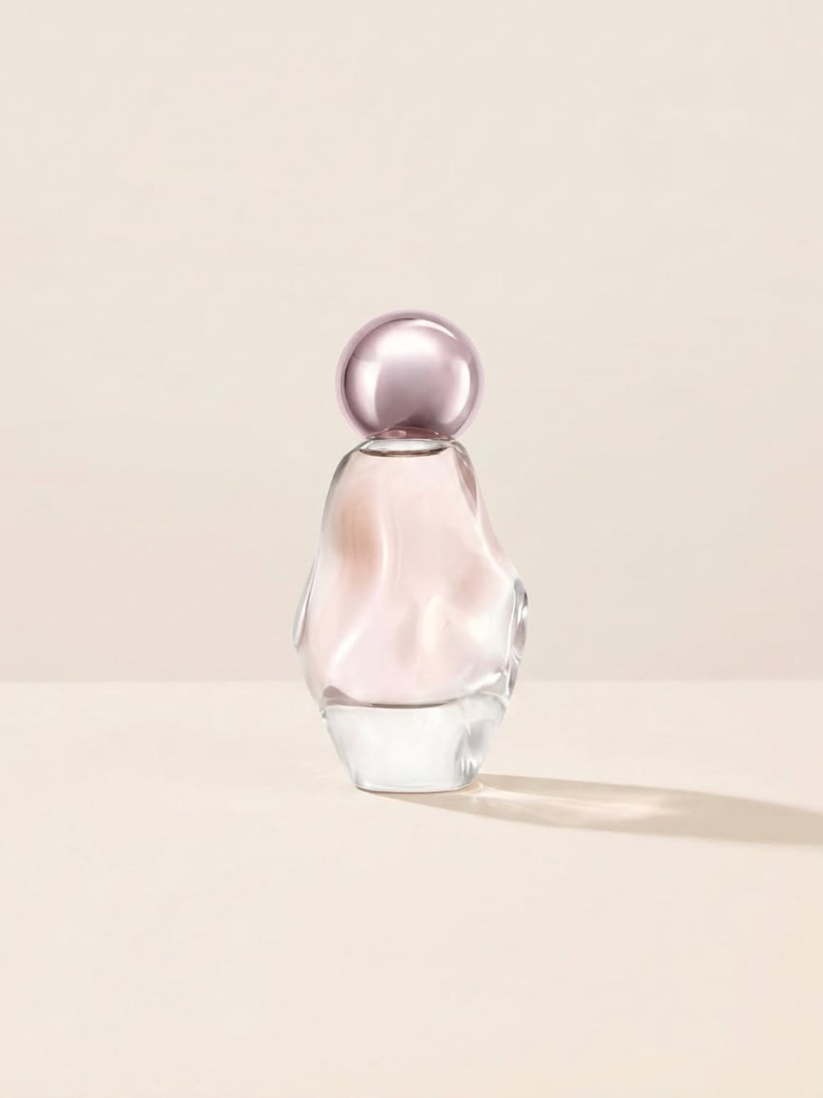 Pear-shaped glass perfume bottle with a lilac sphere cap on a cream backdrop