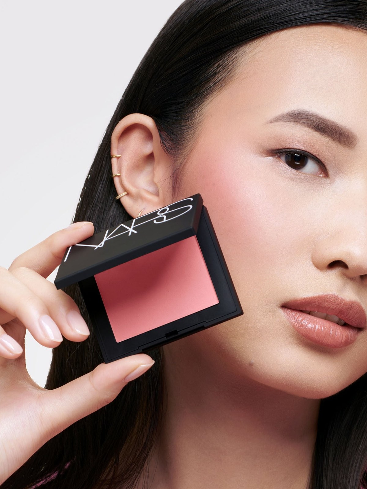 Model with long black hair holding a NARS blush compact beside her face