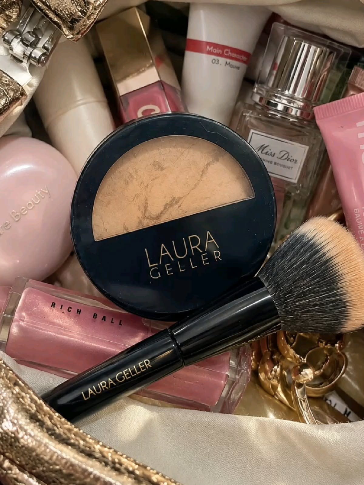Laura Geller bronzer compact and mascara wand nestled among beauty products in a bag