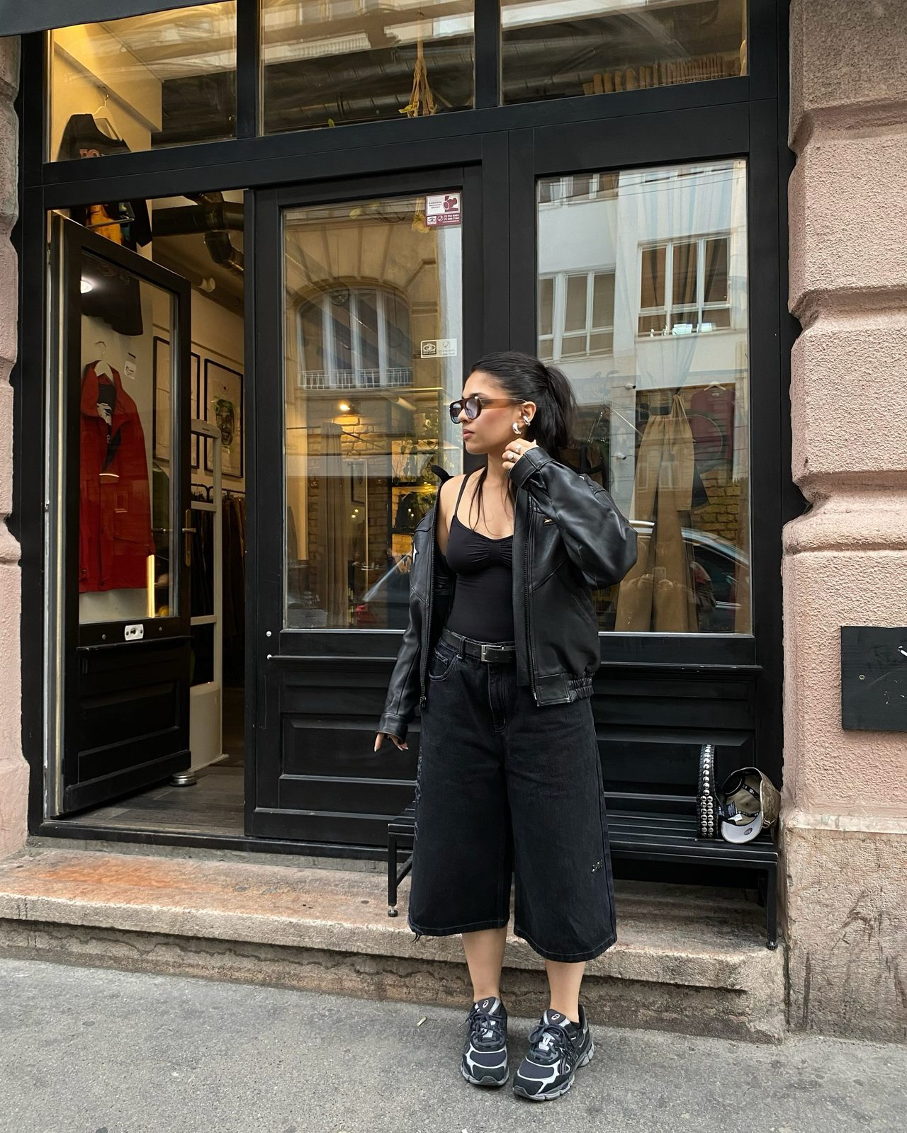 Woman outside a shop doorway in a black top, black leather jacket, black knee-length shorts and black sneakers