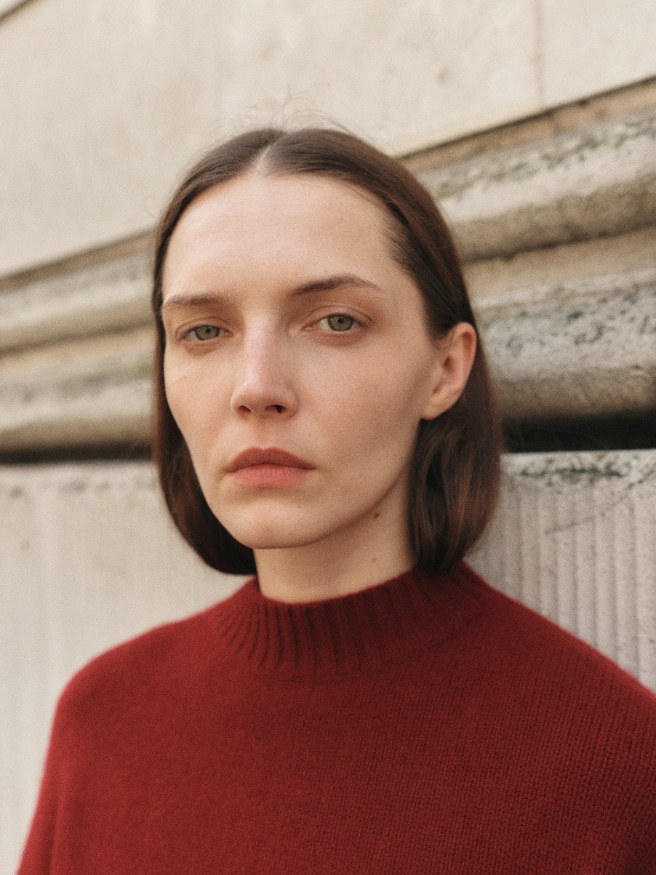 Close portrait of a model in a deep red knit beside pale stone architecture