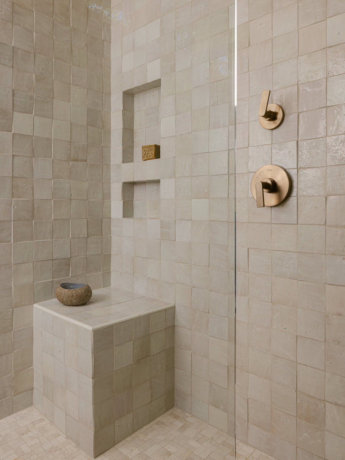 Shower corner tiled in cream handmade zellige tiles with a built-in bench, two niches, a stone bowl and brushed bronze fixtures
