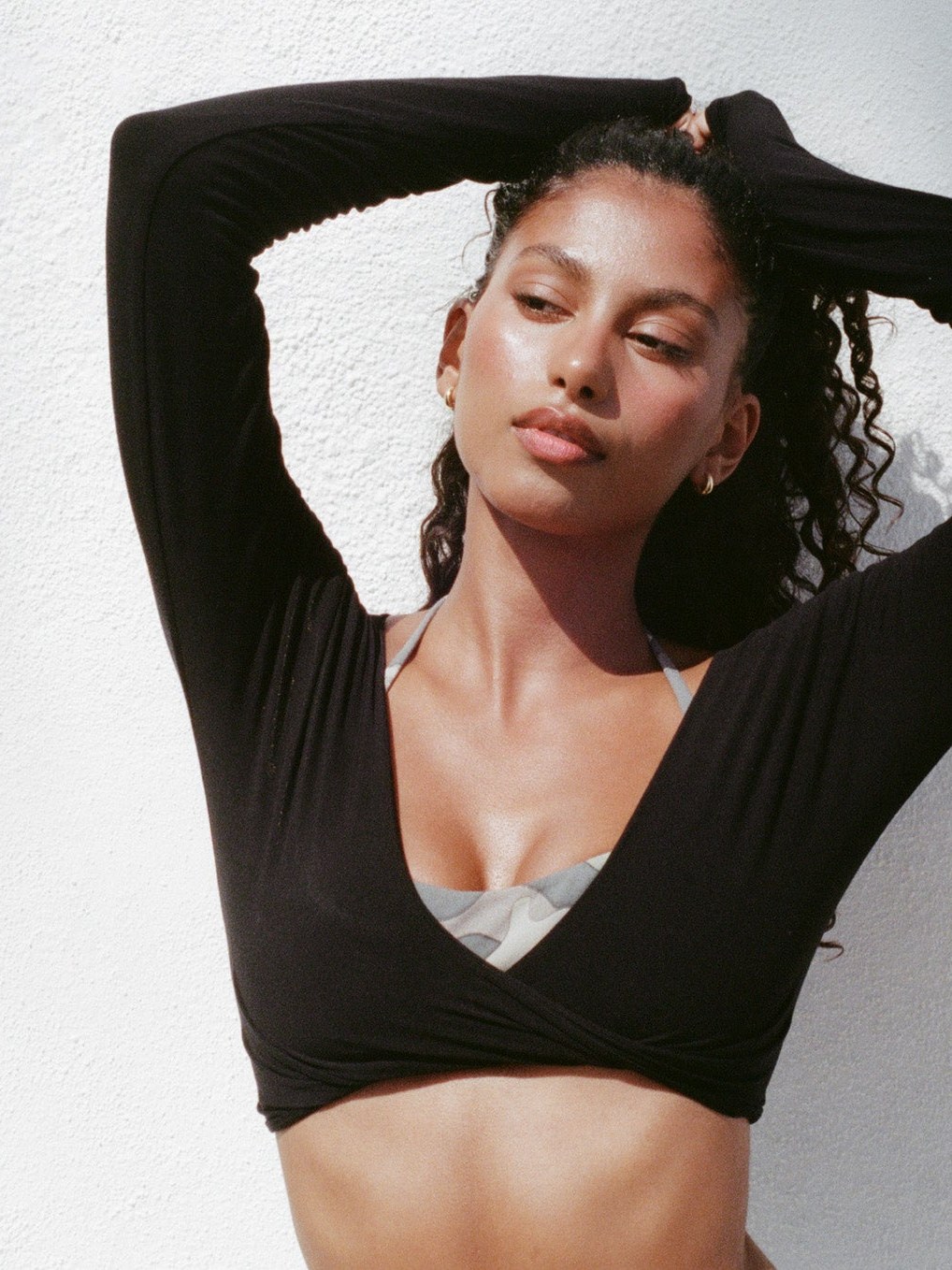 Model in a black wrap top over a light swimsuit against a white wall