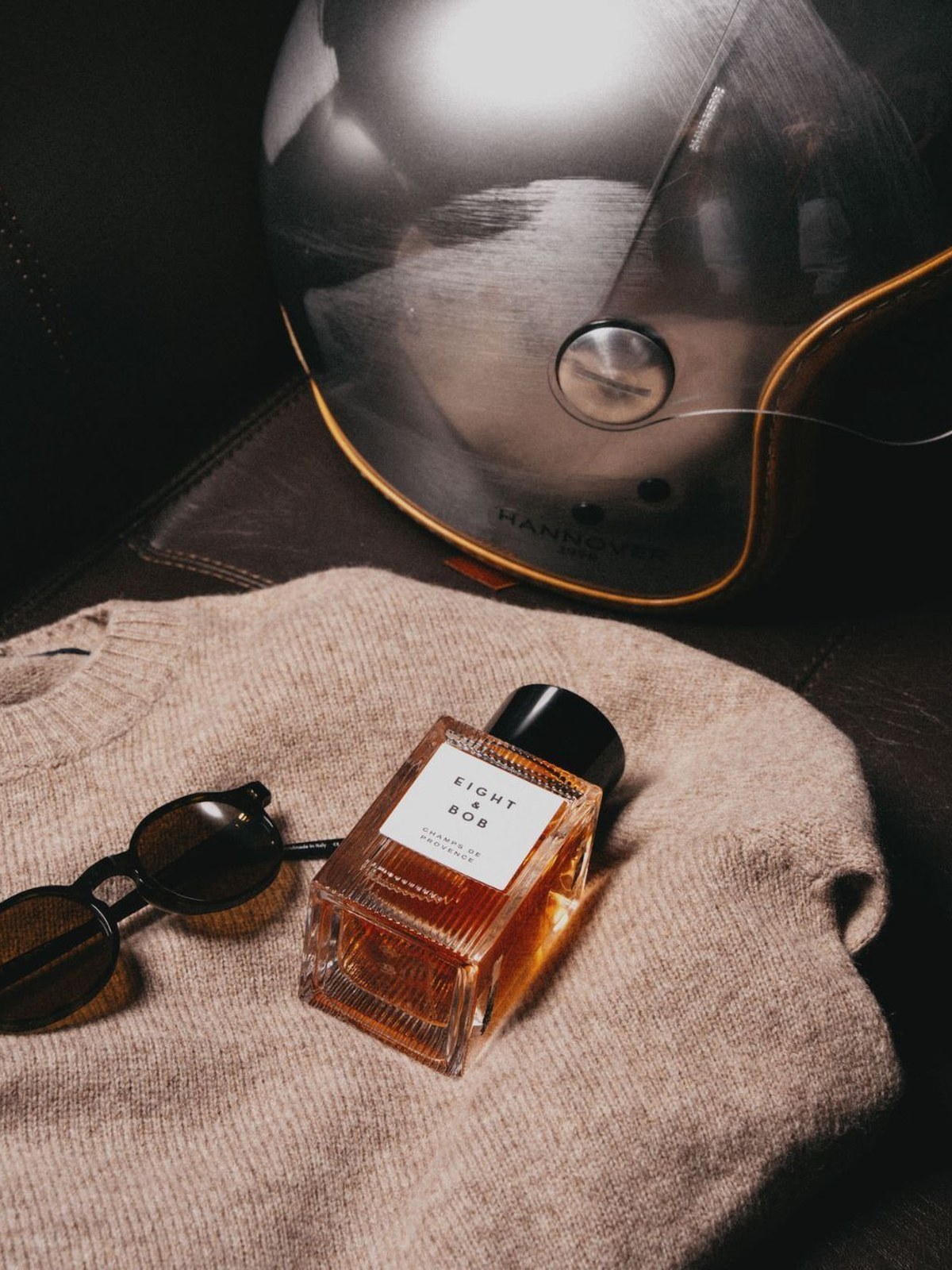 Bottle of Eight & Bob Champs de Provence on a beige knit sweater beside sunglasses and a motorcycle helmet
