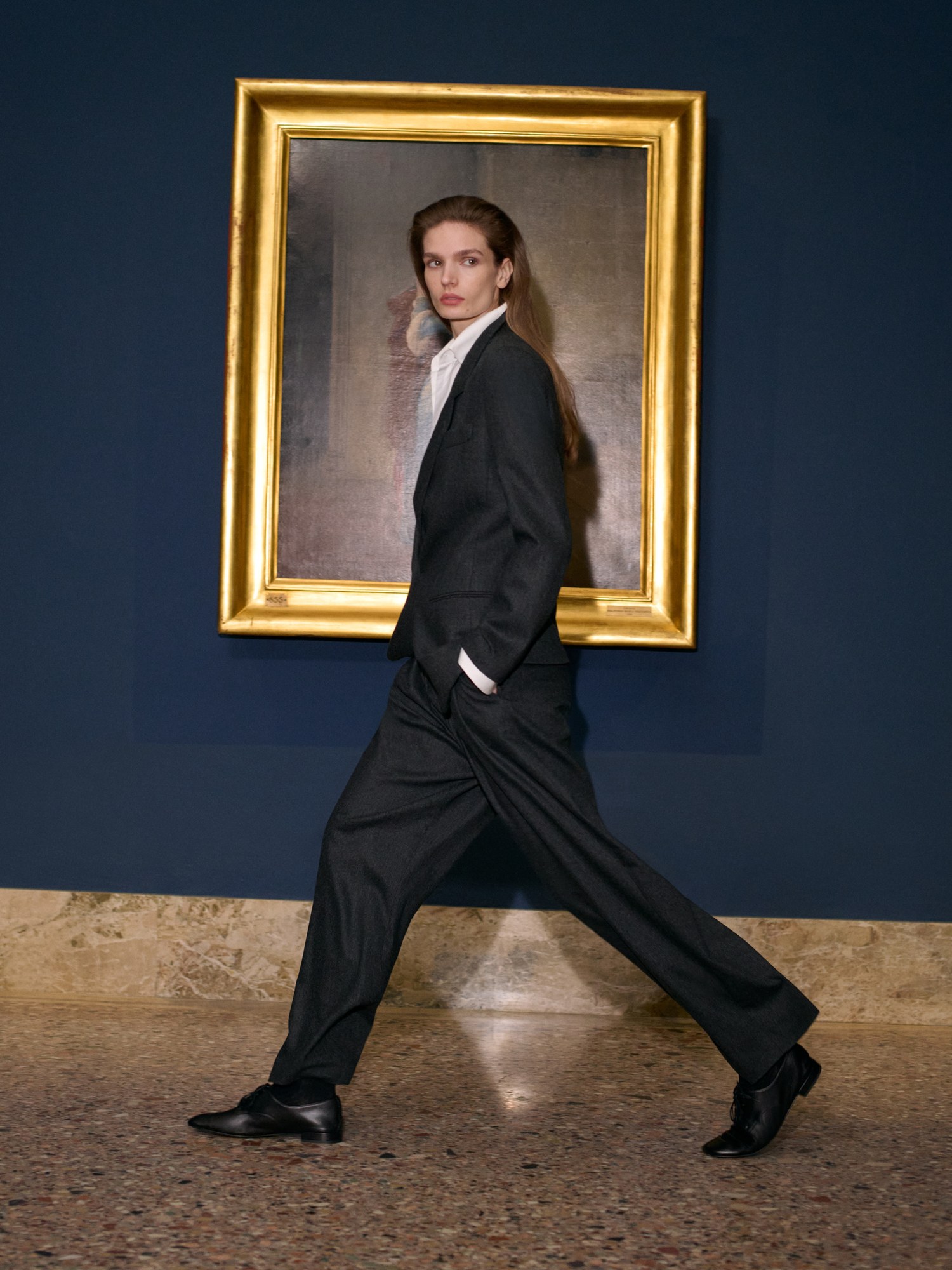 Model striding in a black tailored suit in front of a gold framed portrait