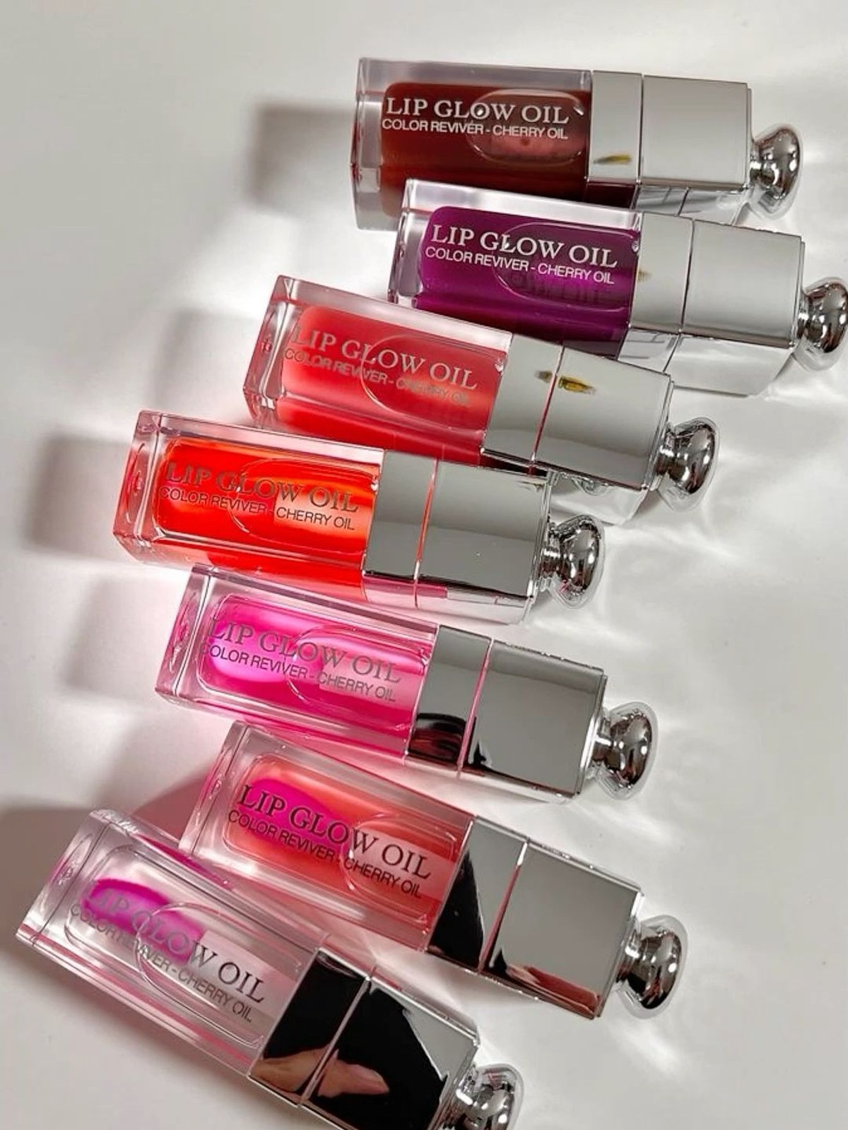Seven Dior Lip Glow Oil bottles lined up on white in shades from clear pink to berry and deep red