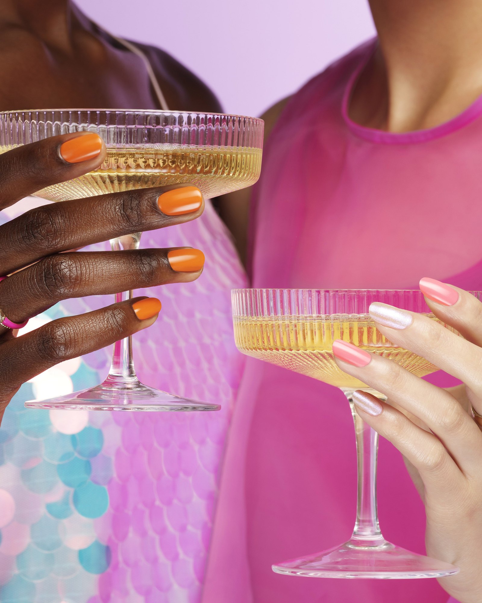 Two pairs of hands holding coupe glasses of sparkling wine, one with orange nails and the other with pink and pearl nails, against a pink sequined background