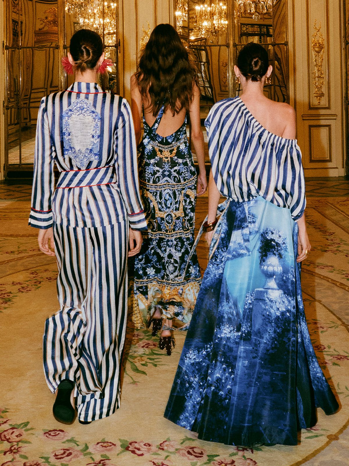 Three models seen from behind in striped and blue printed looks walking through a gilded room
