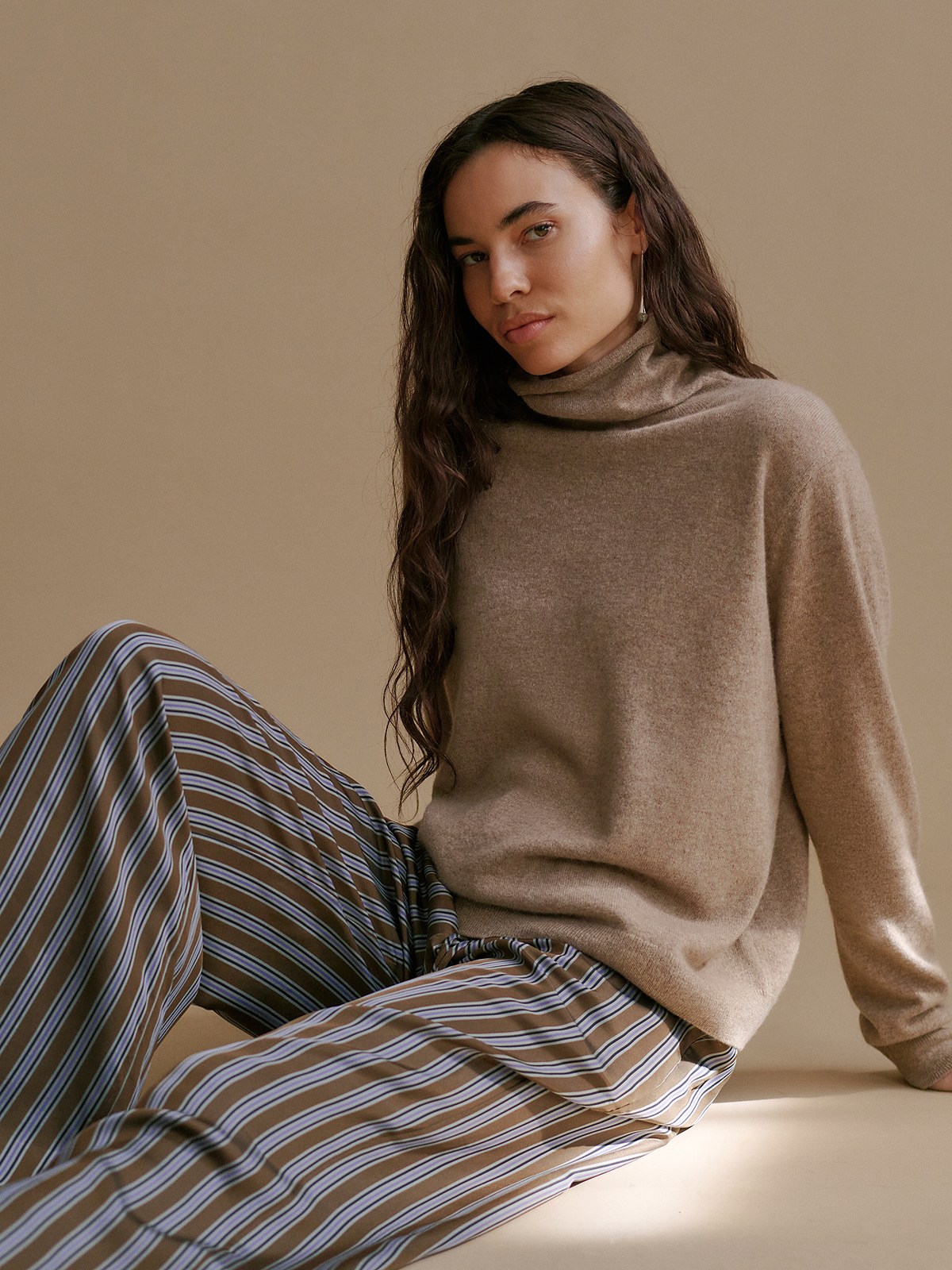 Model seated in an oatmeal cashmere turtleneck and striped wide trousers on a tan backdrop