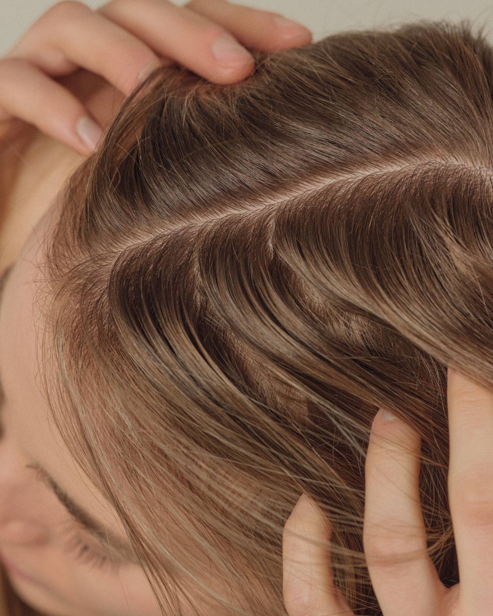 Close-up of a woman parting her light brown hair with both hands to show the scalp