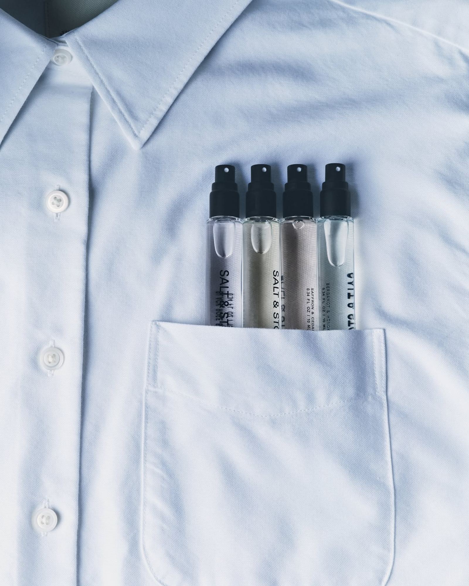 Four small clear perfume atomizers with black caps tucked into the breast pocket of a white button-down shirt