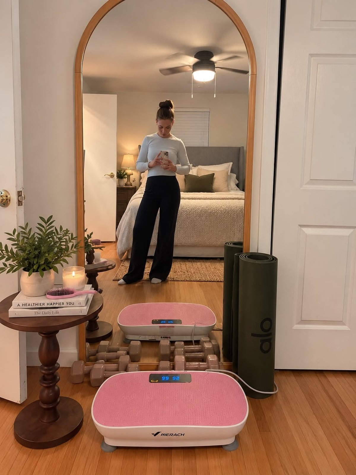 Pink Merach vibration plate on a bedroom floor reflected in a full length mirror next to dumbbells and a yoga mat