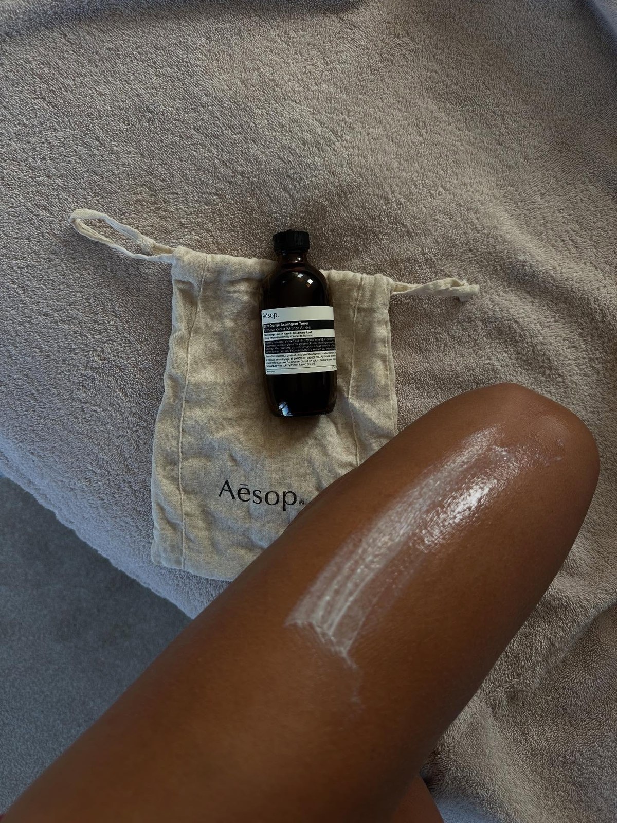 Streak of white lotion smoothed across a bare thigh next to an Aesop toner bottle on a linen pouch and a taupe towel