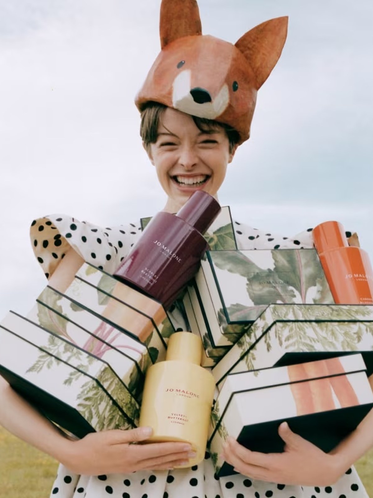 Smiling model in a fox hat and polka dot dress holding an armful of gift boxes in a field