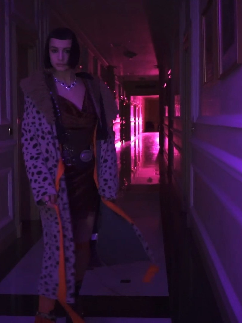 Woman in a spotted coat over a dark dress in a magenta-lit hallway