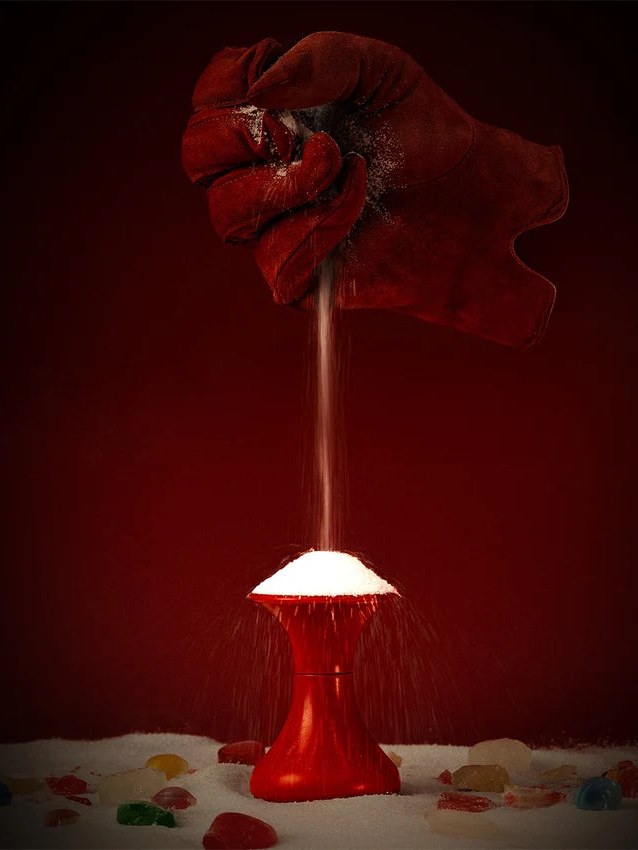 A gloved hand pours sugar over a red bottle in a dark studio.