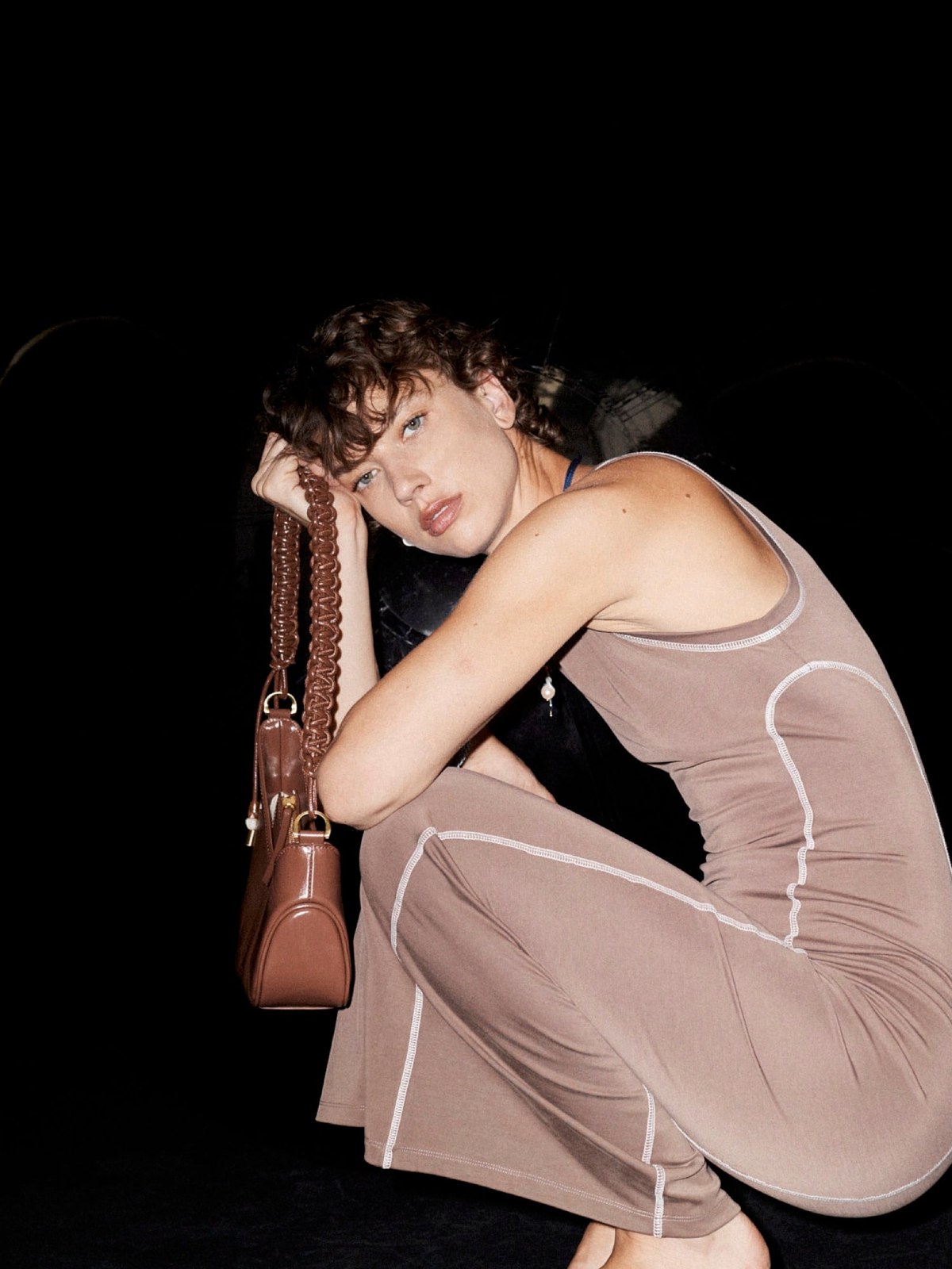 Model in a taupe dress crouching with a brown bag