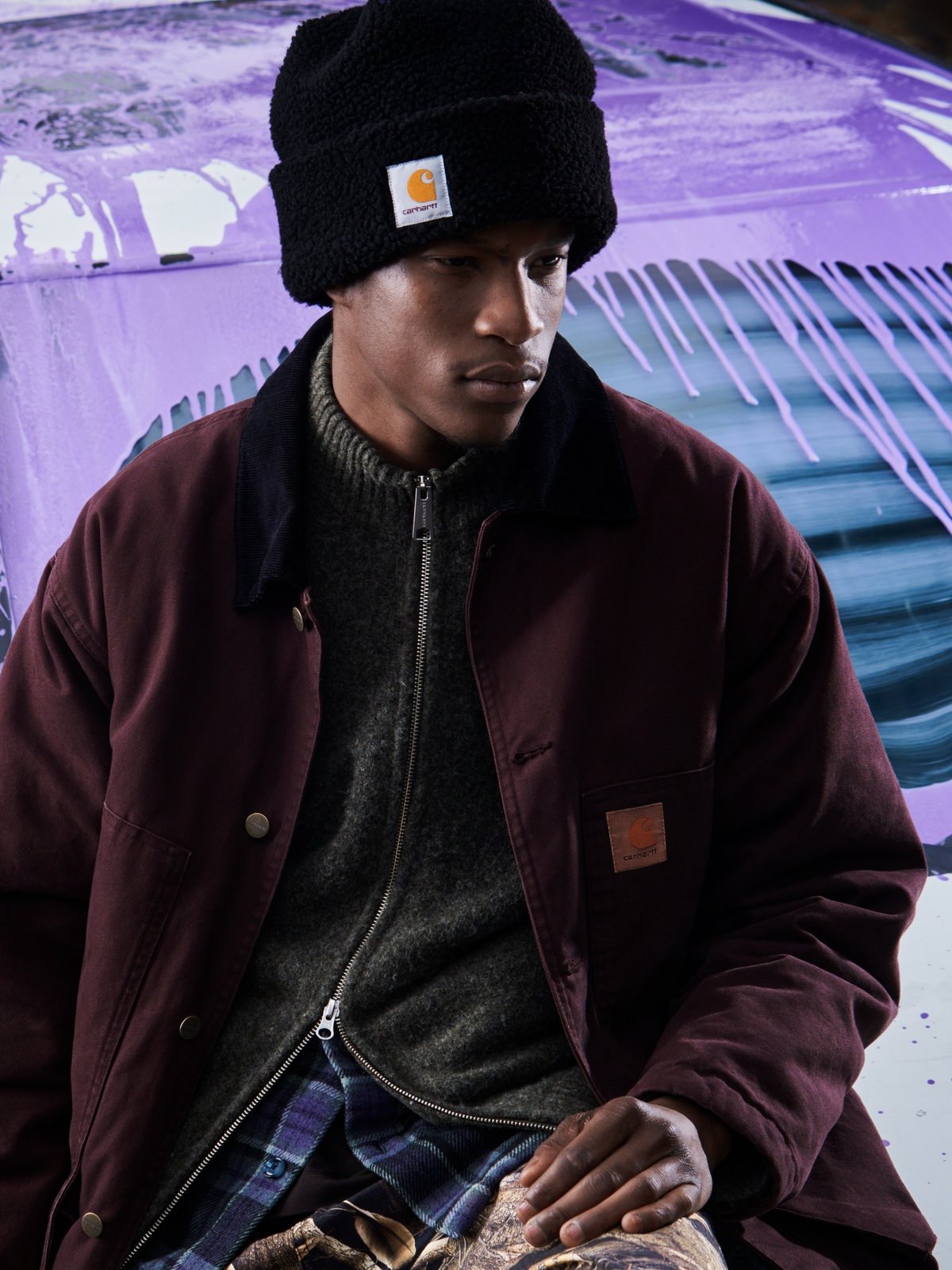 Male model in a black beanie and burgundy chore coat leaning on a paint splattered car