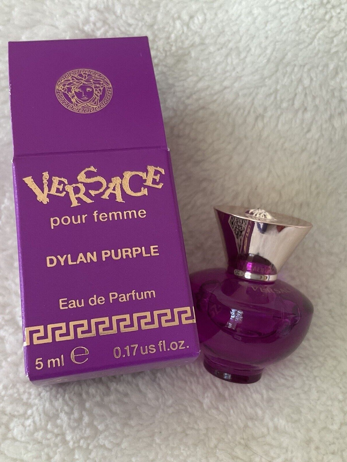 Small Versace pour femme Dylan Purple eau de parfum bottle and its purple gift box with a gold Greek key border on a white fluffy blanket