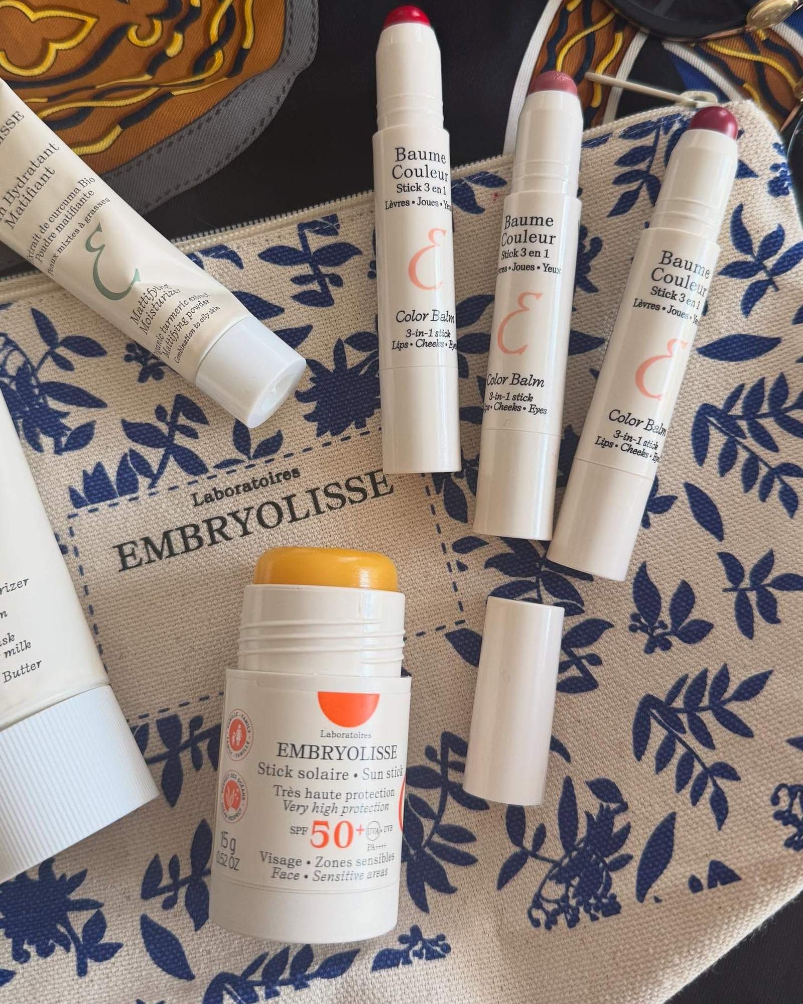 Embryolisse color balm sticks, a sun stick and a mattifying moisturizer tube arranged on a blue and white floral makeup pouch