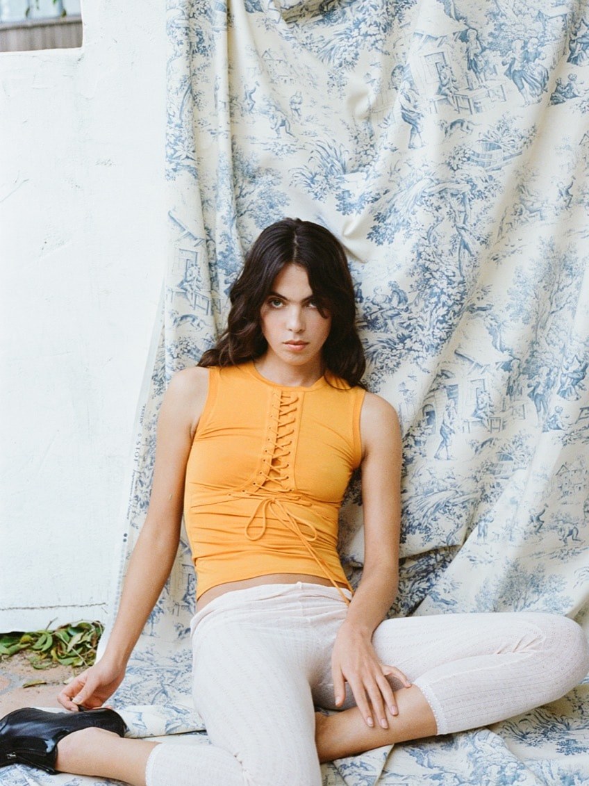 Miaou model seated in an orange top and light trousers against a floral wall.