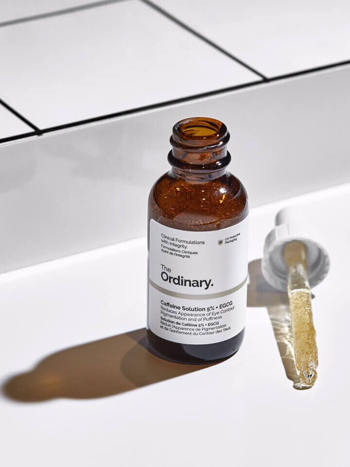 A brown glass bottle of The Ordinary Caffeine Solution 5% + EGCG standing open on a white tiled ledge, its dropper lying beside it in bright sunlight.