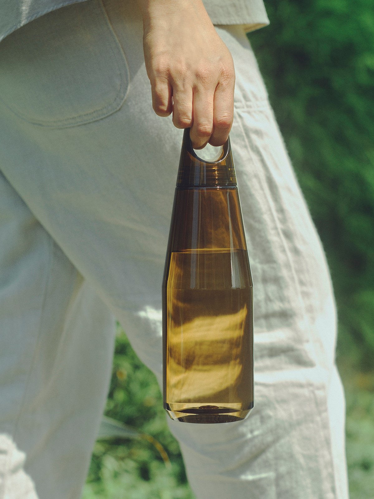 A hand carries an amber bottle against a green garden setting