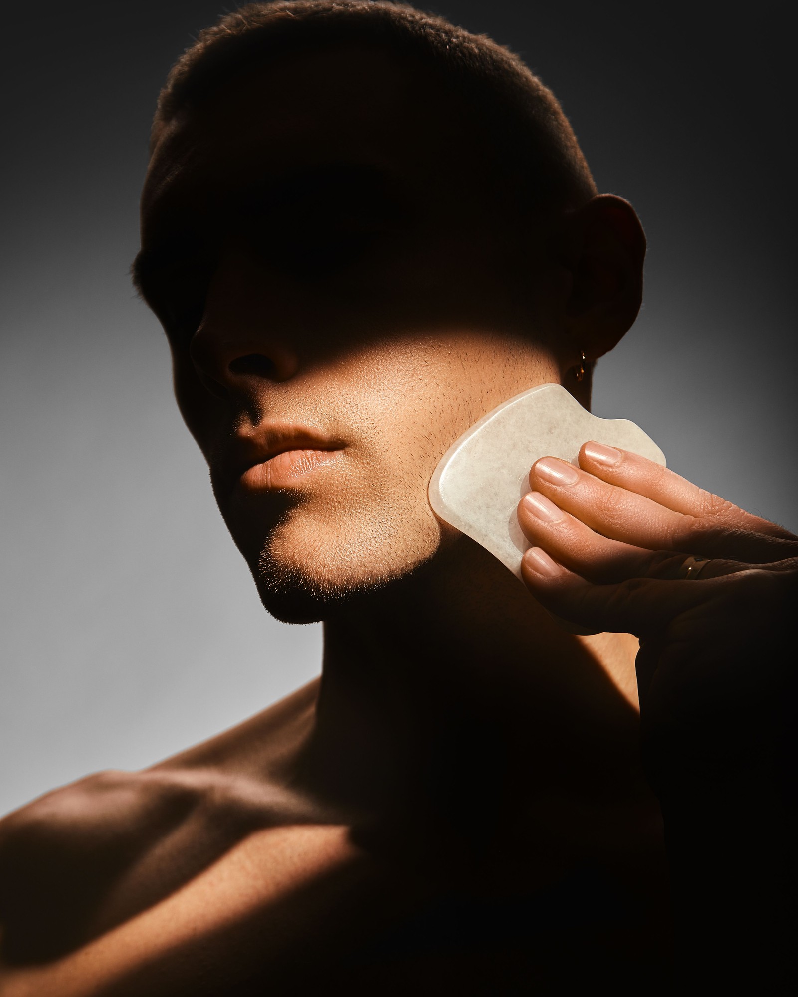 Man in dramatic shadow holding a pale stone facial tool against his jaw
