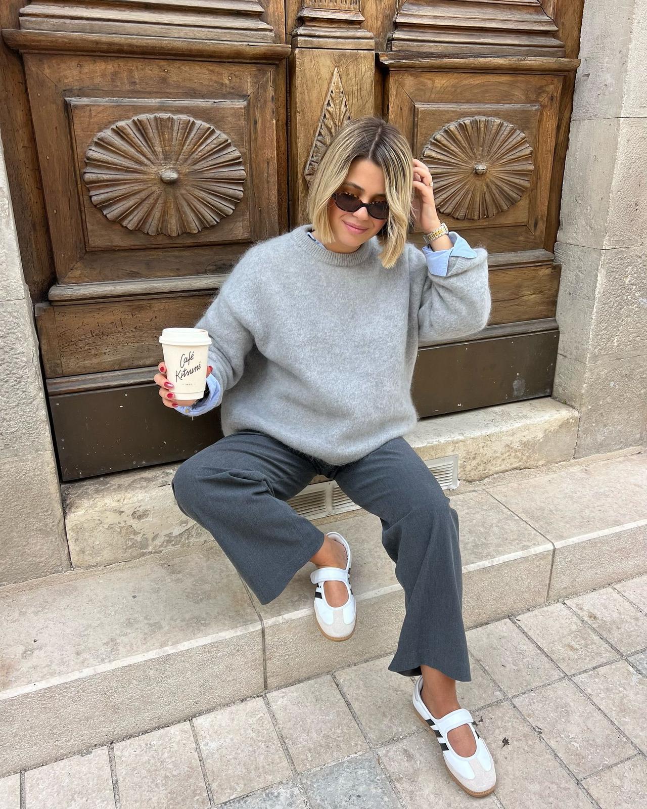 Woman sitting on a stone step in a grey sweater and grey trousers with white Samba Jane sneakers, holding a coffee