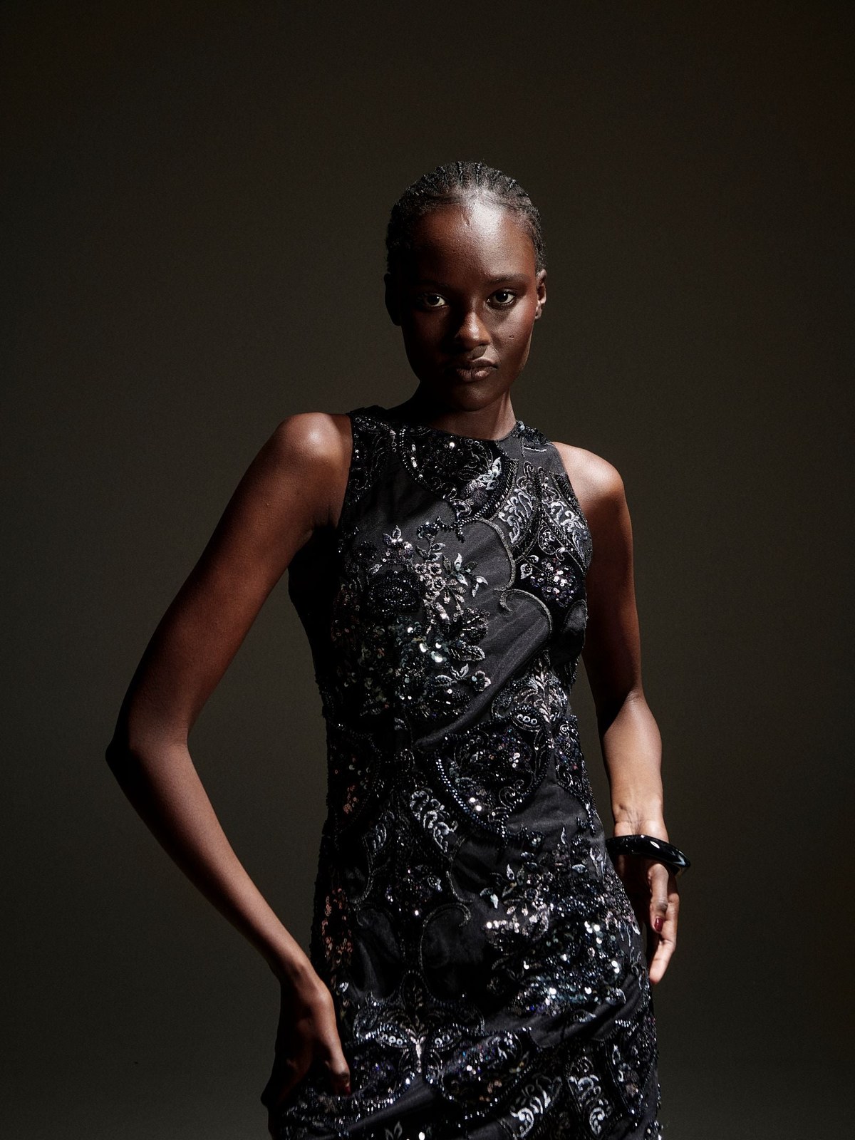 Model in a black embellished sleeveless dress against a dark backdrop