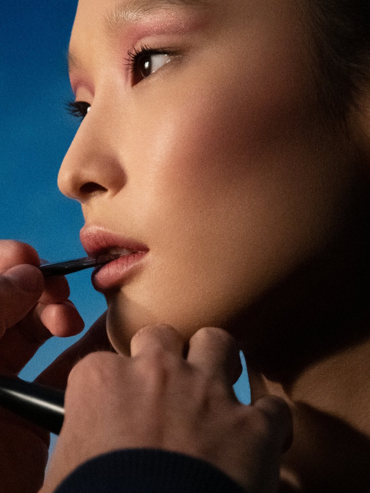 Model in profile having lip colour applied with a brush against a blue backdrop