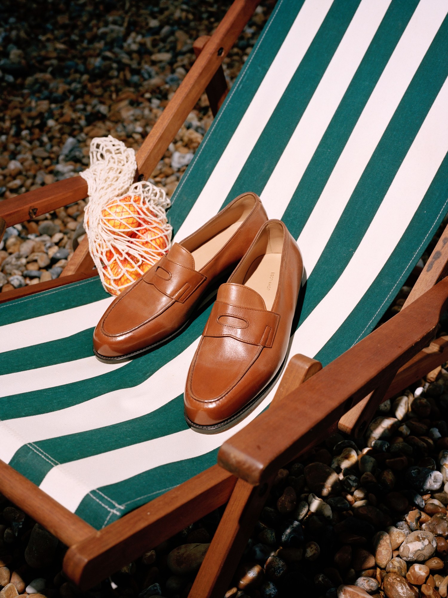 Tan penny loafers on a green striped deckchair on a pebble beach