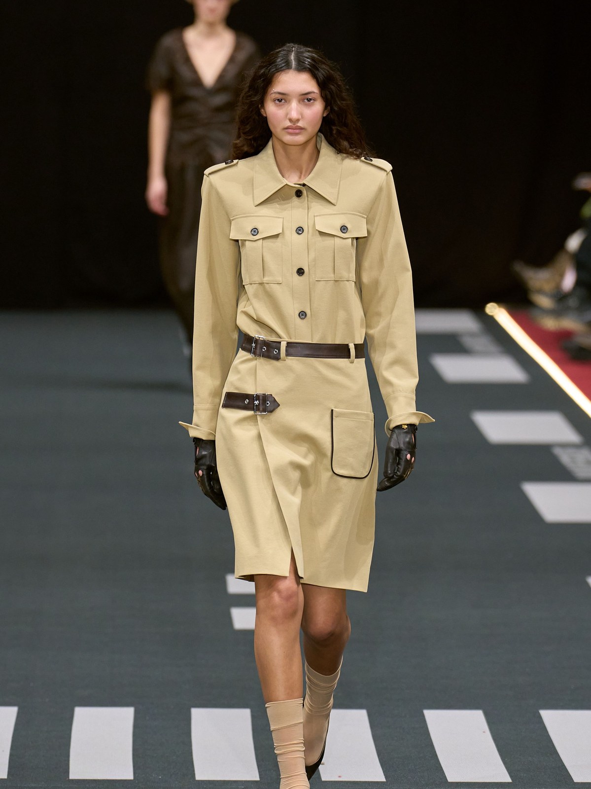 Runway model in a light beige belted coat and dark gloves
