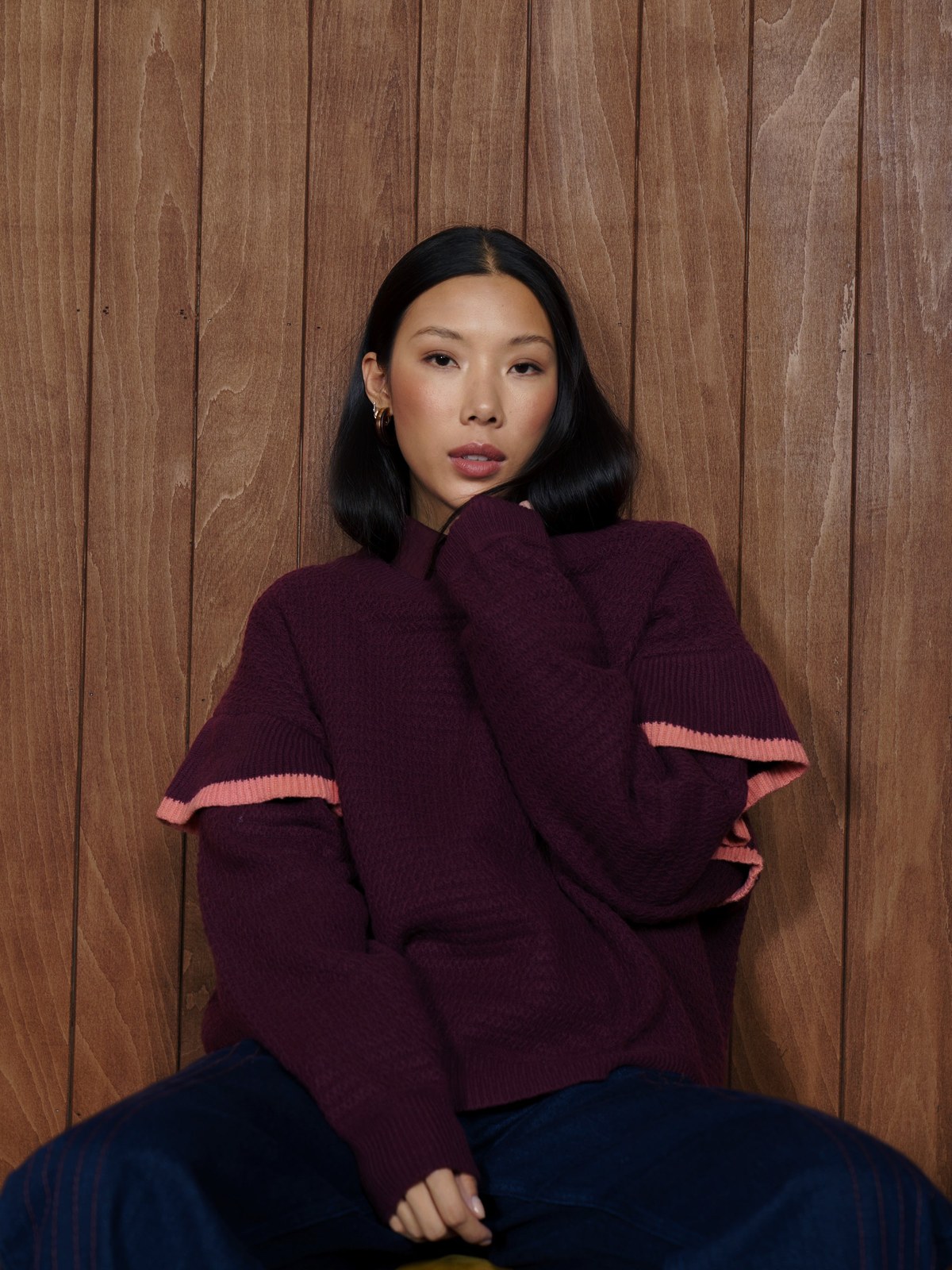 Model seated in a burgundy sweater with dark wide-leg jeans against a wood wall